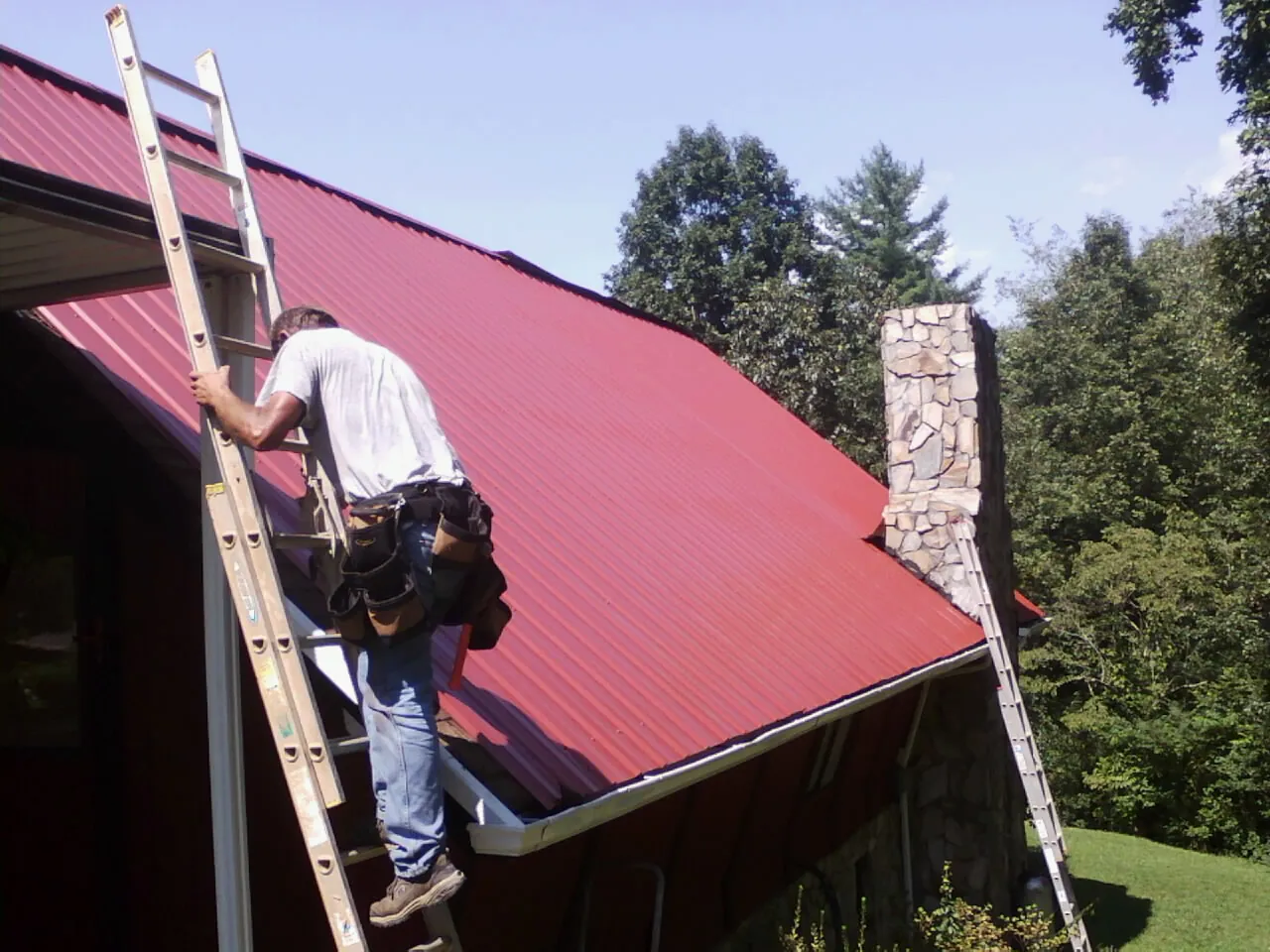 Roofer on a ladder beside a red metal roof and stone chimney — Soffit & Fascia Repair by Roofing Brothers in Marble Falls